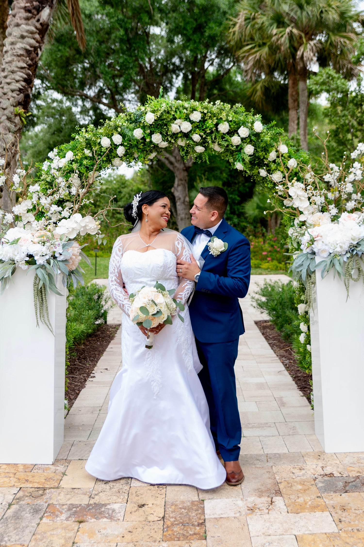 A bride in a white lace wedding gown, design by Alexas, holding a bouquet of white and green flowers stands next to a groom in a navy blue suit with a white boutonniere, walking under a lush green and white floral archway on a stone pathway.