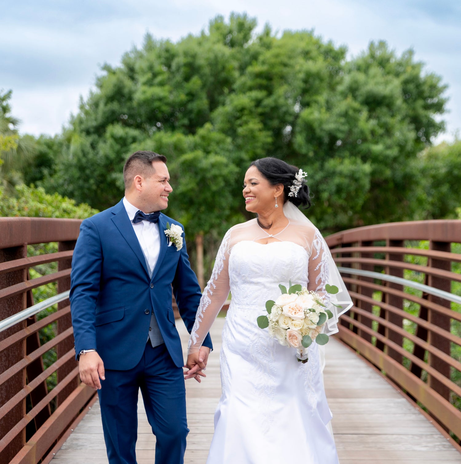 A bride in a white lace wedding gown holding a bouquet of cream and pink flowers walks hand in hand with a groom in a navy blue suit on a wooden bridge with green trees in the background.