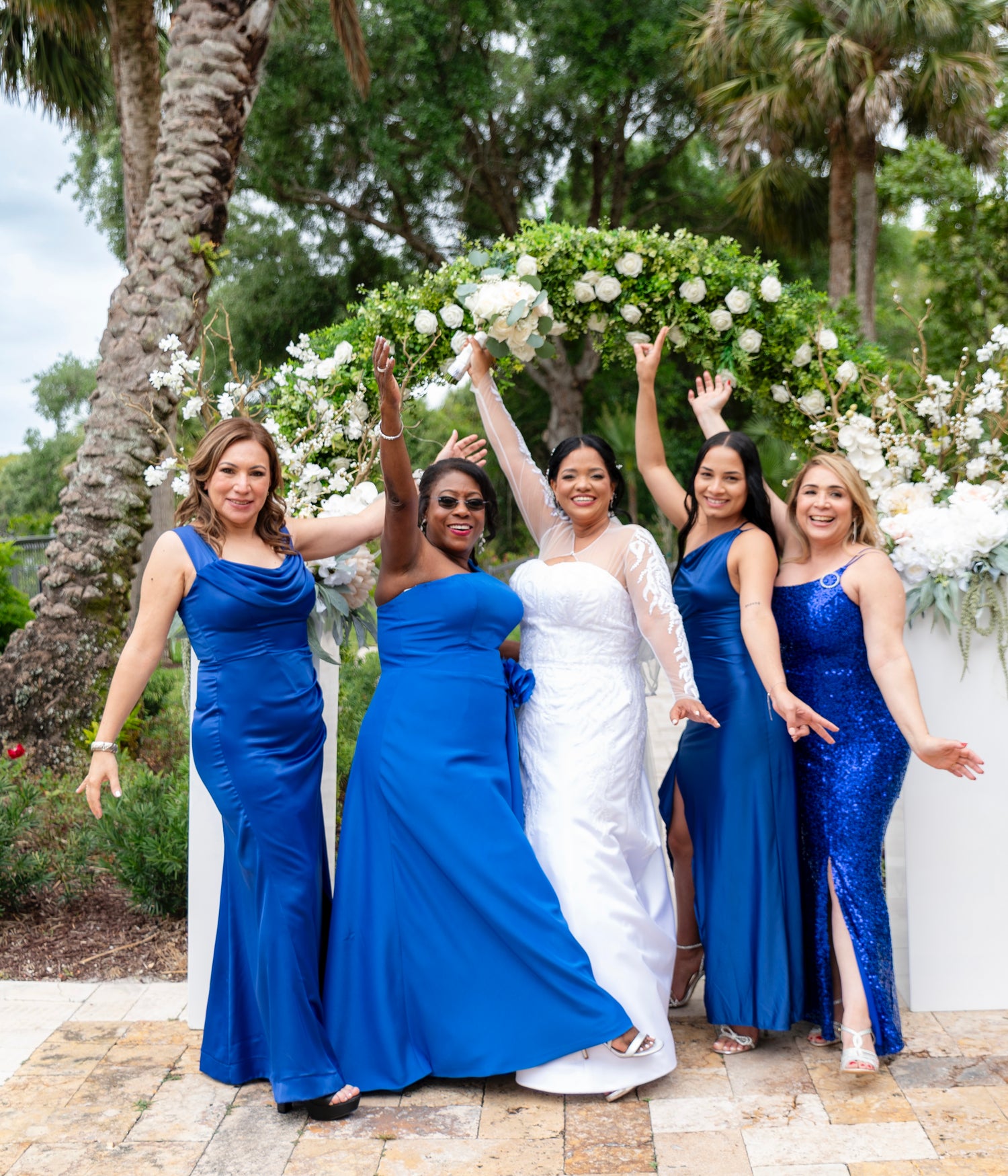 Five women posing together outdoors in front of a white floral arch. Four women wear floor-length blue bridesmaid dresses, design by Alexas