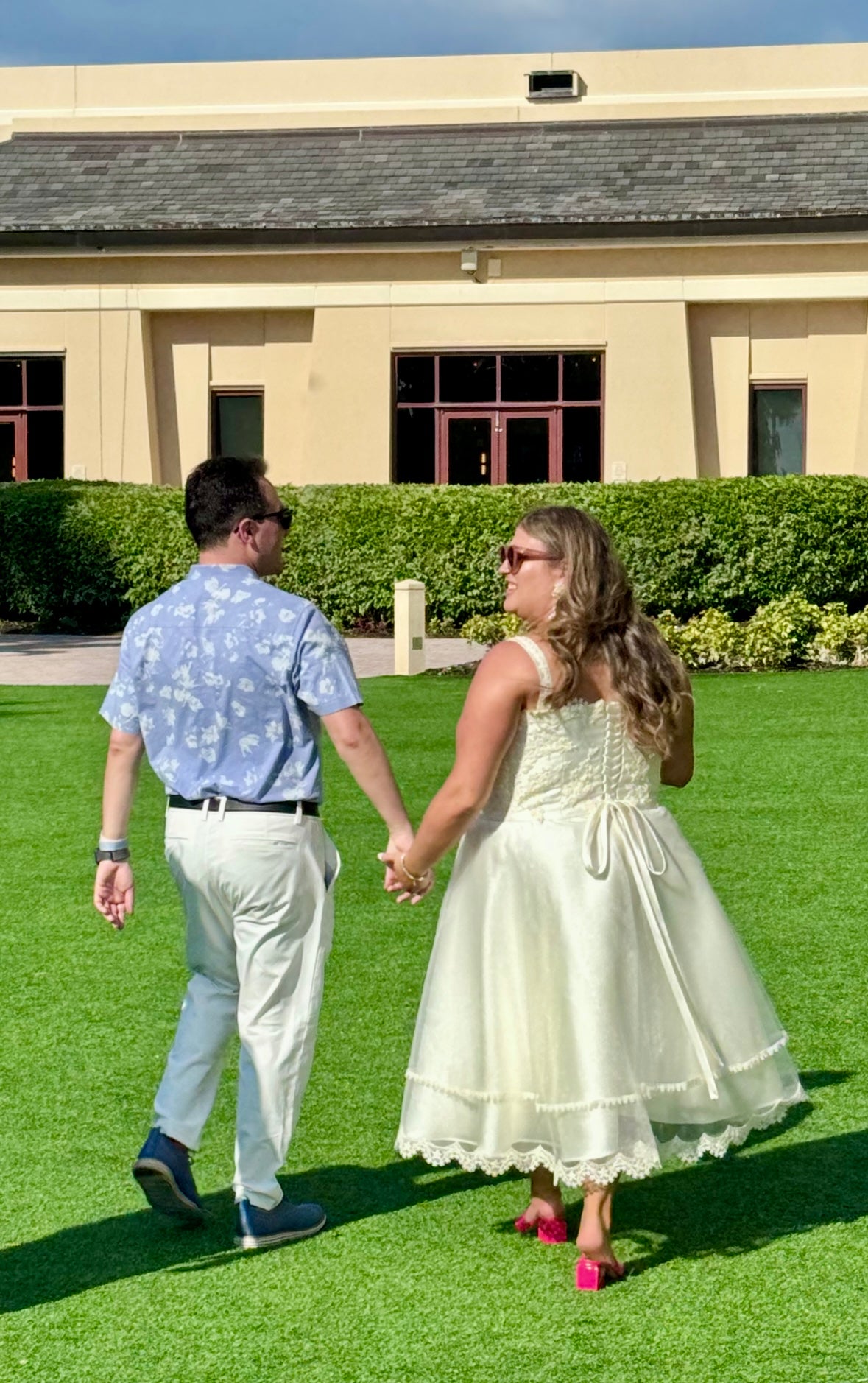 A couple holding hands while walking on a green lawn in front of a beige building. The woman is wearing a cream-colored sleeveless dress with a lace-trimmed hem and a bow at the back, design by Alexas