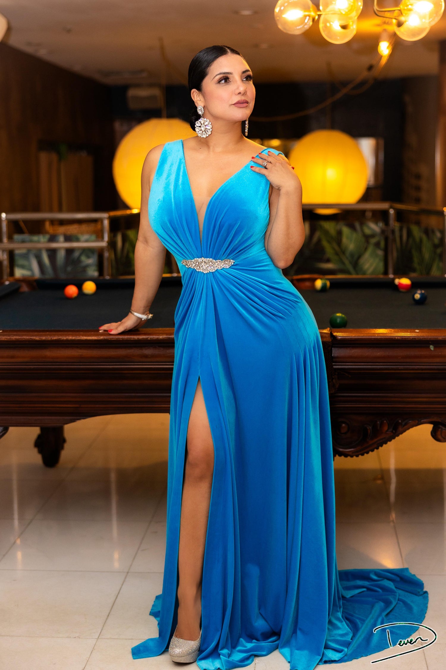 A woman wearing a vibrant blue floor-length gown with a plunging neckline, a high thigh slit, and a draped waist detail, design by Alexas