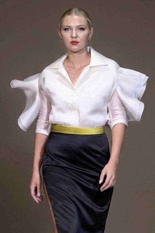 A model wearing a white long-sleeve blouse with dramatic ruffled sleeves and a black satin skirt, design by Alexas