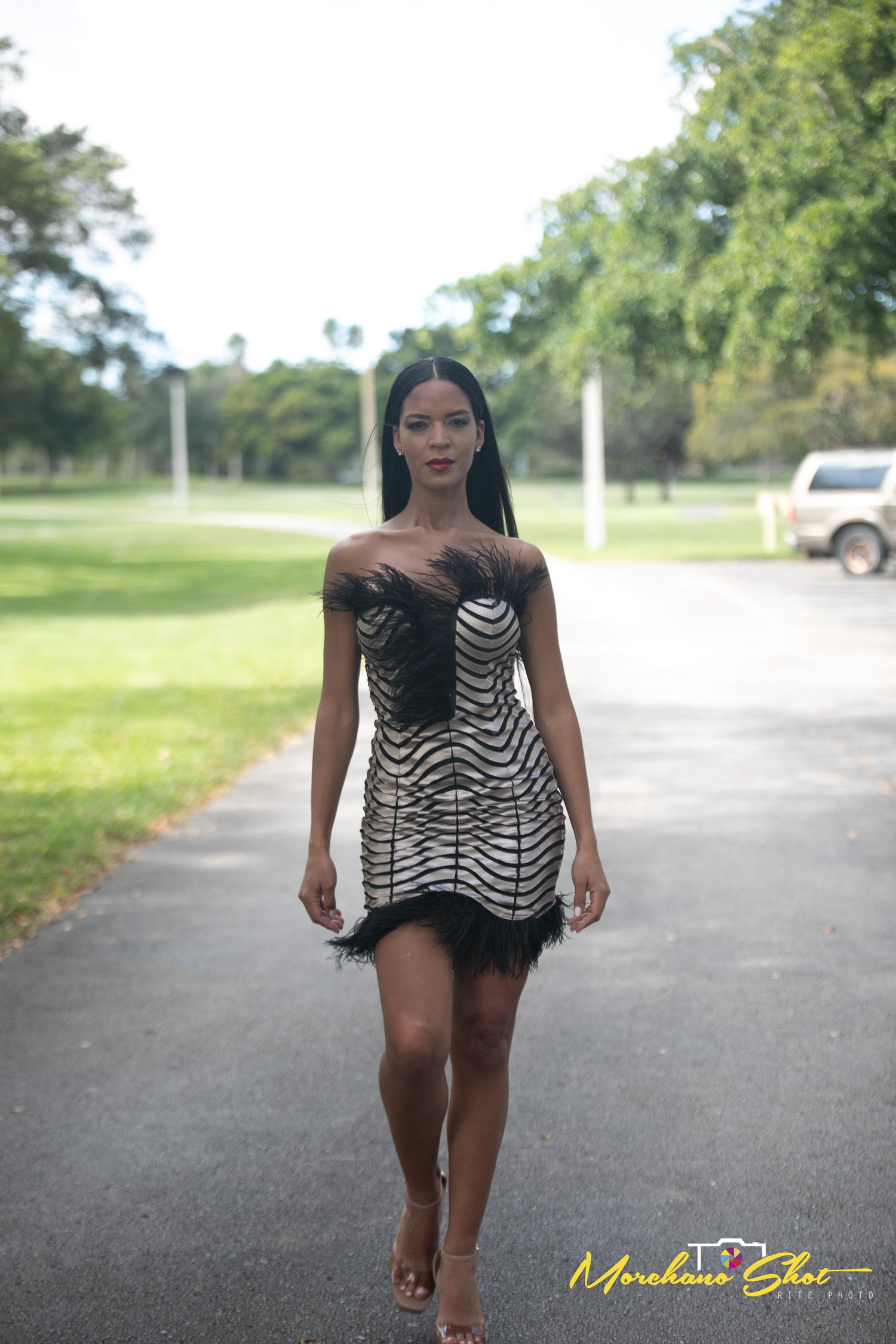 A woman walking on an outdoor paved path wearing a strapless black and white striped mini dress with black feather-like trim at the bust and hem. Design by Alexas