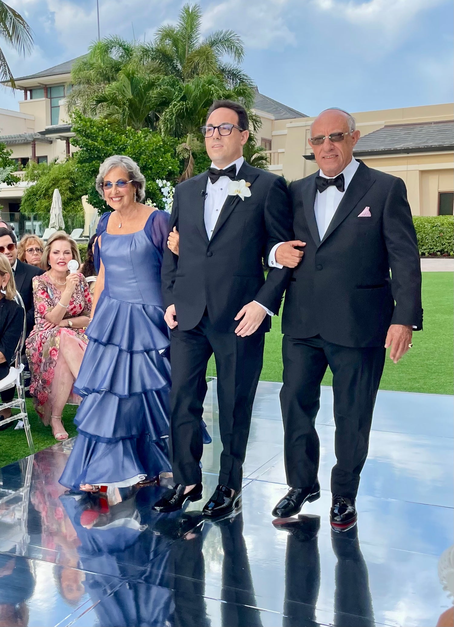 Three adults walking outdoors on a reflective black surface, with two men in black tuxedos and a woman in a blue tiered gown, design by Alexas