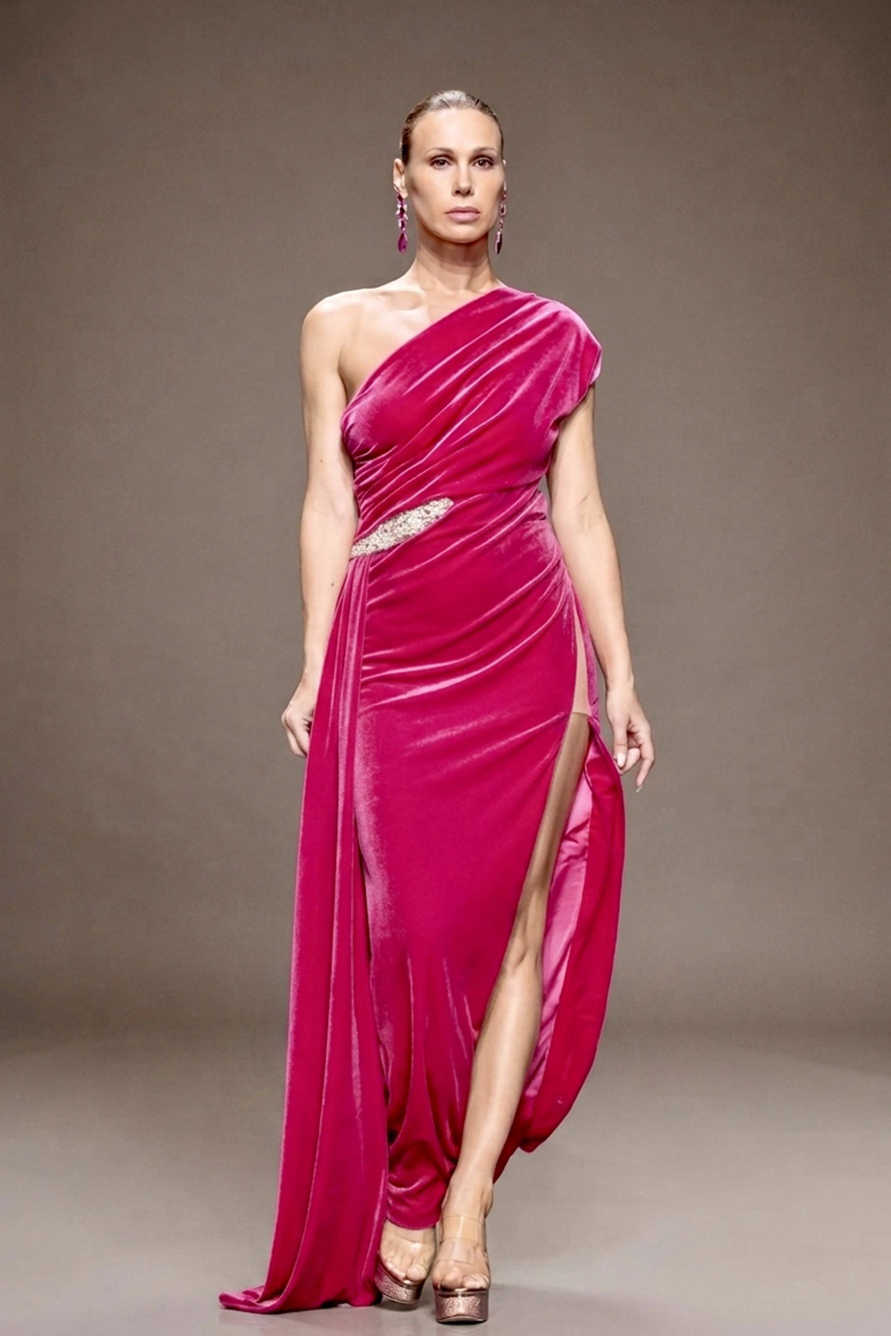 A woman modeling a floor-length draped gown in vivid fuchsia pink velvet. The one-shoulder dress features a high thigh slit and a sparkling embellished waist detail, design by Alexas