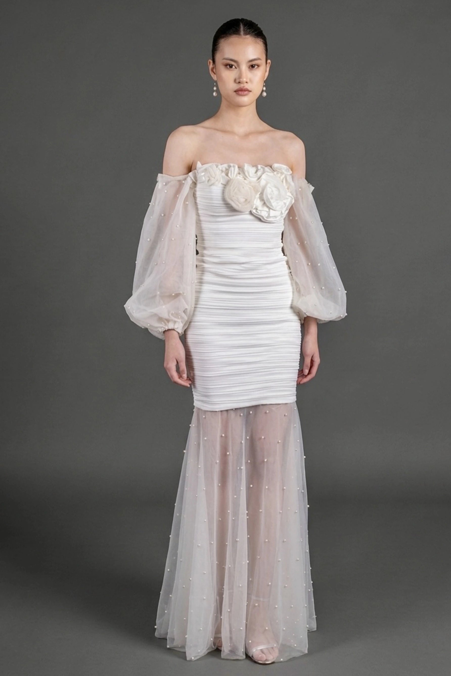 A model wearing an off-the-shoulder white gown with voluminous sheer puff sleeves, a textured ruched bodice with large floral applique, and a sheer tulle mermaid skirt with subtle bead details, design by Alexas