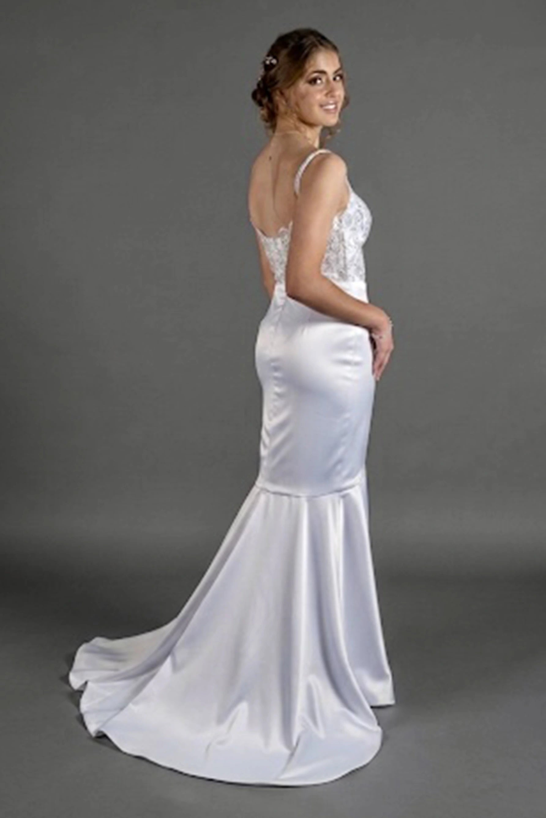 A woman modeling a white satin mermaid-style wedding dress with a lace bodice and backless open back, design by Alexas