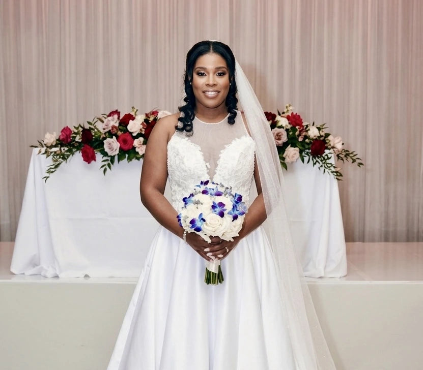 Sarah in a white wedding dress by Alexas and veil holding a bouquet of white and purple flowers, standing in front of a decorated backdrop with floral arrangements.