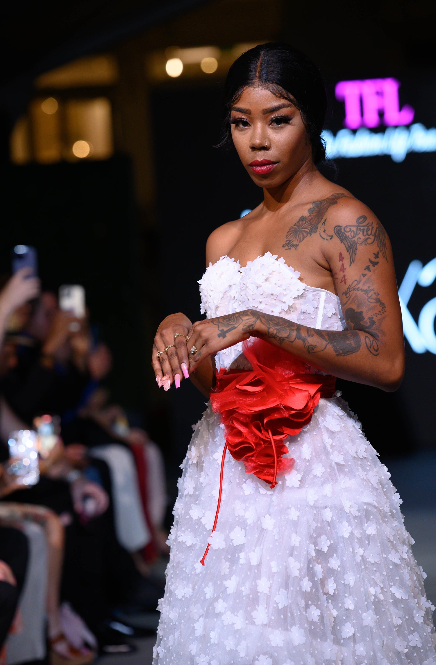 A model walks a fashion runway wearing a strapless white textured dress with a prominent red ruffled waist sash. Design by Alexas