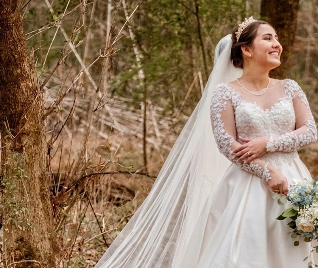 A woman wearing a white lace wedding dress with a long sheer veil, standing outdoors in a wooded setting and holding a bouquet of blue and white flowers.