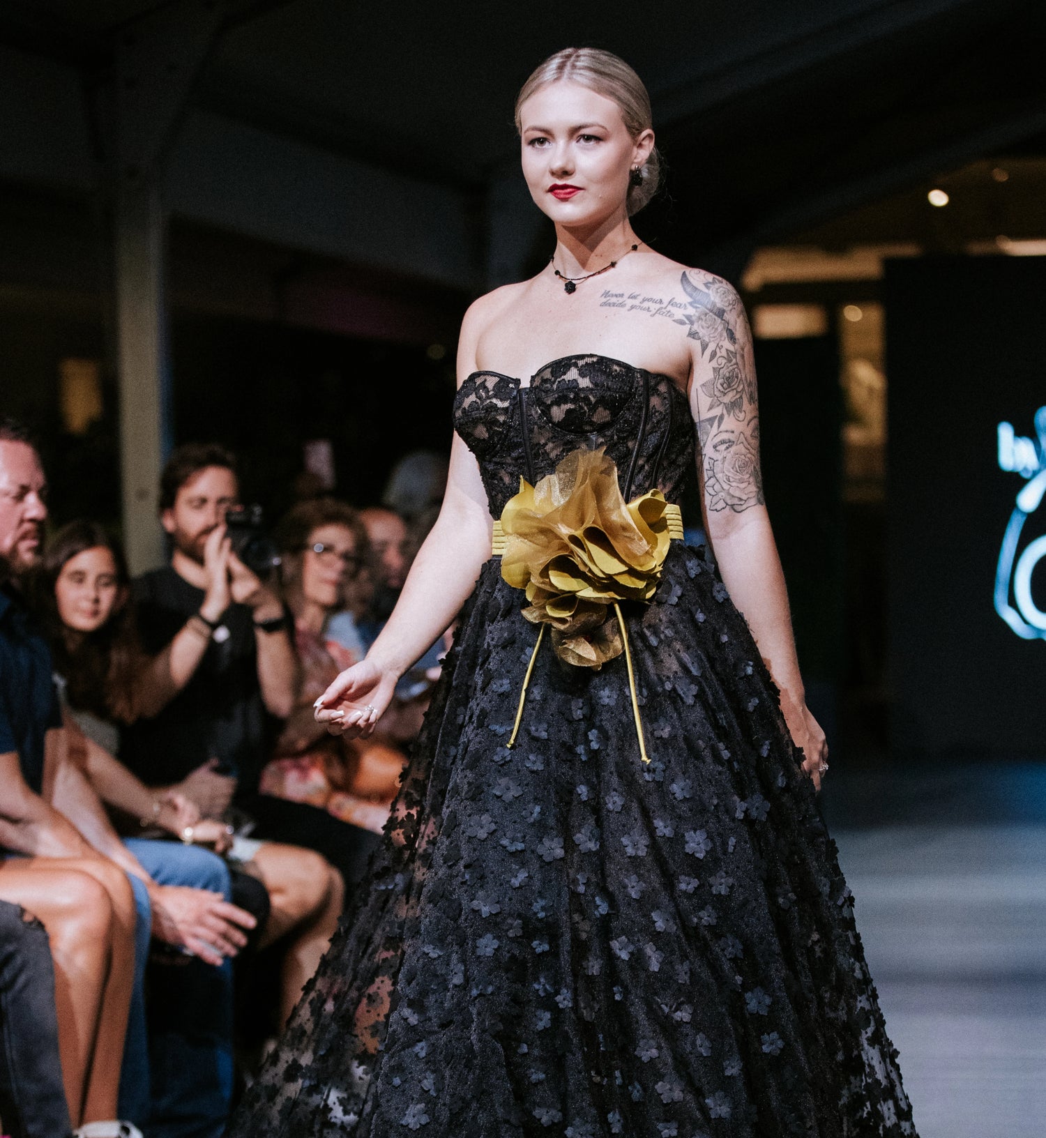 A model walks a fashion runway wearing a strapless black lace ballgown with a large gold fabric flower at the waist, design by Alexas